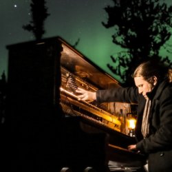 A man sits at an upright piano underneath an immersive projection of the aurora borealis