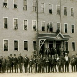 Black and white photo of a large wooden factory building with a line of men standing if front of it posing for the camera.