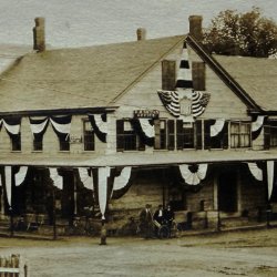 Black and white photo showing the corner of a village street with two and three story buildings festooned in patriotic bunting.