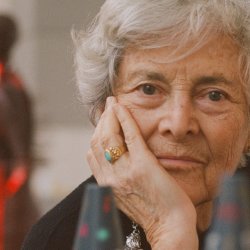 A woman with short grey hair looks at the camera with her head resting on her hand. Around her are cone shaped sculptures, some in and some out of focus.