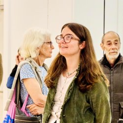 People gathered in a white walled gallery looking at the artworks. In the centre is a person with glasses wearing a green jacket looking up at a textile sculpture.