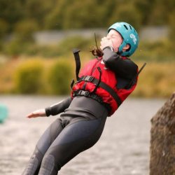 Young person wearing a green helmet and red life jacket leaning back and falling into the water during an outdoor activity session.