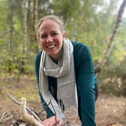 Hannah is smiling at the camera. She has her hair pulled back, is wearing a dark green jumper and a pale grey scarf is wrapped loosely round her neck. She is standing a wood leaning on a pile of stick