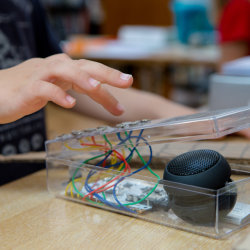 Close-up of a hand interacting with a small electronics project in a clear plastic container, containing coloured wires, components and a speaker. The image represents hands-on digital making, technol