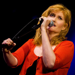 A woman holding a microphone with a red cardigan and black top on with shoulder length hair
