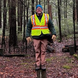 A man in a safety vest stands perched on two pieces of wood stuck in the ground in the woods. The wood appears to be part of a an old foundation or structure.