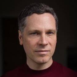 Colour head and shoulders picture of Allan Radcliffe wearing a burgundy top and standing in front of a darkened blurred room