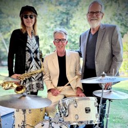 Three Trio Metro musicians outdoors behind a drum kit: a woman in sunglasses and a beret holding a saxophone, a smiling man in a cream suit holding drumsticks, and a bearded man in a gray blazer.