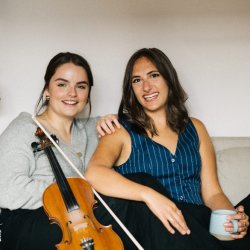 Rachel Campbell (right) and Holli Scott (left) are sitting facing the camera and smiling. Rachel is resting her hand on Holli's shoulder. A fiddle rests on Rachel's body.