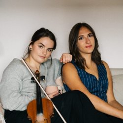 Rachel Campbell (right) and Holli Scott (left) are sitting facing the camera and smiling. Rachel is resting her hand on Holli's shoulder. A fiddle rests on Rachel's body.