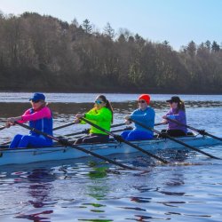 Four people in vibrant athletic gear row a boat across a calm, sunlit river.