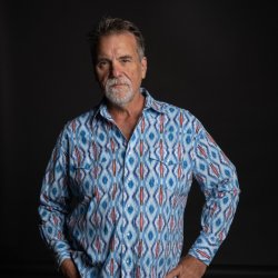 Darrell Scott standing against a dark background wearing a patterned blue shirt