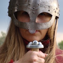 A close up photo of a long haired woman wearing an Anglo-Saxon iron helmet with eyepiece and nose protection and holding up a sword so the hilt of the sword is visible.