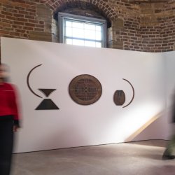 A blur of people move past a pink and black piece of art hung on a red brick wall