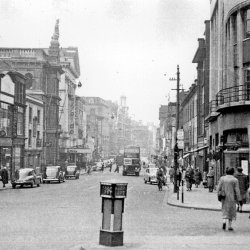 A black and white photograph of Granby Street, Leicester. There are people walking on the pavement and cars parked along the street. In the middle of the road in the foreground is a Keep Left bollard.