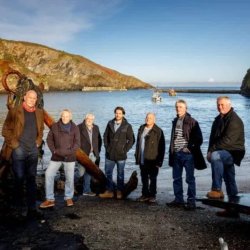 Seven men are stood with jeans and jackets in front of a sea cove scene
