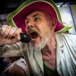 A man with a goatee wearing a wide-brimmed pink and green hat sings into a microphone.