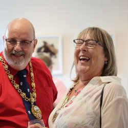 The Mayor and Mayoress of New Romney share a laugh at a JAM private view, framed artwork displayed on the wall behind them.