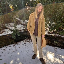 Cam Norton, long blonde hair, in a tan corduroy jacket and pants holding a hat, standing outdoors in front of green foliage.