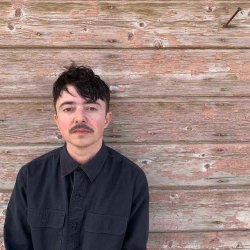 Dean Spade, a white trans person with short straight dark brown hair and a mustache stands in front of a wooden wall. He wears a dark grey button up shirt.