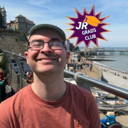 Jack, a white man with black glasses, smiles to camera. He wears an orange t-shirt. A beach is in the background.