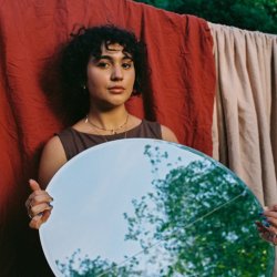 SUNNY is in a brown dress, standing in front of a red sheet with a mushroom beige sheet layered next to it. she is holding a circular mirror reflecting a tree and blue sky ahead. Curly brown hair