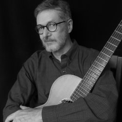 Singer-songwriter Pierce Pettis sits holding an acoustic guitar, looking toward the camera in a black-and-white portrait.