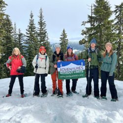 6 people wearing snowshoes pose for a photo with the A-W-L flag on a snowy, tree-lined trail