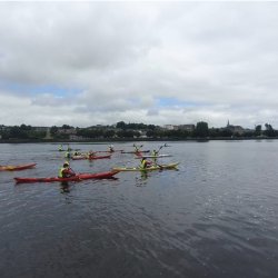 A group of kayakers wearing high-visibility vests paddle colourful kayaks across a wide river under a cloudy sky, with a town and shoreline visible in the distance.