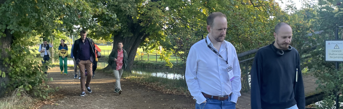 A group of people walking along a footpath in pairs. There are green trees overhanging the footpath. Two men are walking in front.