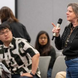 Image shows a women speaking into a microphone. She is in a large room with other people watching her speak.