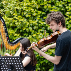 James Warburton (violin, front) and Angharad Huw (harp, back) performing outside with a big leafy bush as a backdrop. They are both wearing black t-shirts and looking at their music.