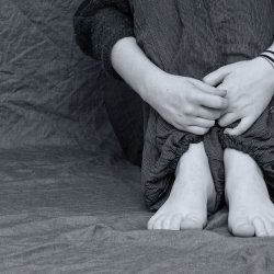 someone, possibly a teenager, is sat against a wall - knees pressed to the chest and arms around their shins