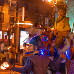 A juggler leads a glowing fish with rider through a nighttime street parade