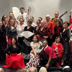 A densely-packed group of Spanish dancers dressed in red, black and white, holding aloft flamenco fans, standing alongside a couple of flamenco guitarists.