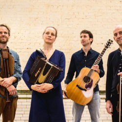 The image features Naragonia Quartet standing in front of a cream painted brick wall holding their instruments
