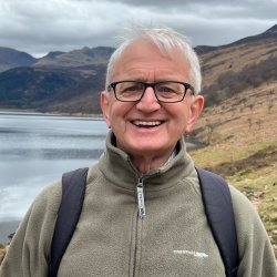 Colour head and shoulders picture of Roger Crofts standing in a winter glen with hilside and a loch in the background. He is wearing a beige zipped up fleece and carrying a rucksack.