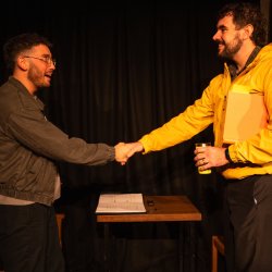 Two men shake hands across a small table during a performance of The Borrowed Room.