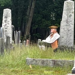 Man in a tricorn hat, vest and breeches seated at the base of a gravestone gazing into the distance.