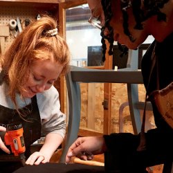 2 people are in a workshop setting, the one on the right is directing the other person how to upholster a dining chair seat with black fabric, using an air powered staple gun.