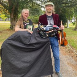 The image features Jennifer Reid and Tom Kitching standing in a tree lined path behind a black, covered, skeletal horse. Tom is holding a fiddle. in his left hand.