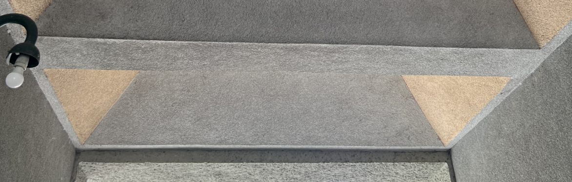 Ein Hauseingang, der beton in ocker und grau gestrichen, links eine Lampe ohne Lampenschirm