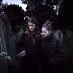 two girls standing infront of a gravestone in a dark churchyard and reading the text on the stone.
