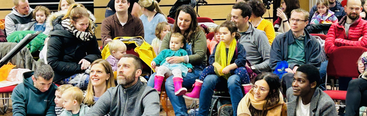 Audience members - parents with babies and toddlers. Some sitting on chairs, others on the floor.