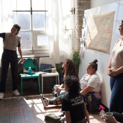 A person standing in front of a bright sunny window points at a whiteboard, sharing their work, while others sit and stand around them.