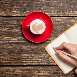wooden background with a red cup and a heart in the foam, there is a notebook with a hand writing on the left