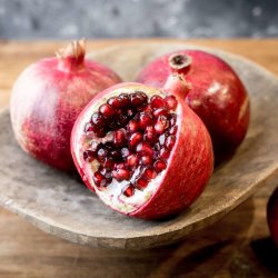 Three pomegranates on a wooden plate. One pomegranate has a wedge removed revealing the seeds inside.