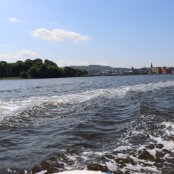 Boat wake rippling across the River Foyle under a clear sky.