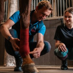 A coach and student crouch next to a horse, the coach showing how to apply a bandage to the horse's front leg