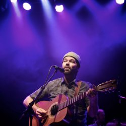 Musician Joel Van Horne of Covenhoven performs onstage with an acoustic guitar under purple and blue concert lights, singing into a microphone during a live performance.